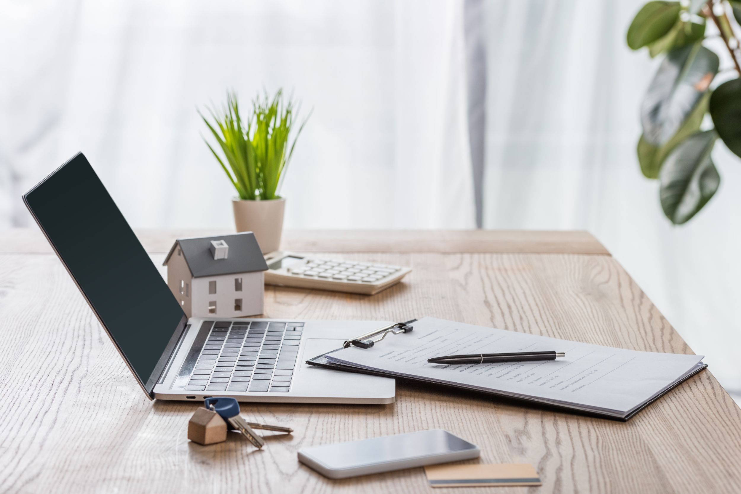 Wooden desk with laptop, smartphone, house keys, clipboard with pen, house model and green plant Wooden desk with laptop, smartphone, house keys, clipboard with pen, house model and green plant
